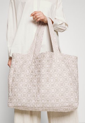 Personne portant une chemise blanche tient un grand sac cabas matelassé rose clair avec un motif géométrique en cercle et des poignées larges.