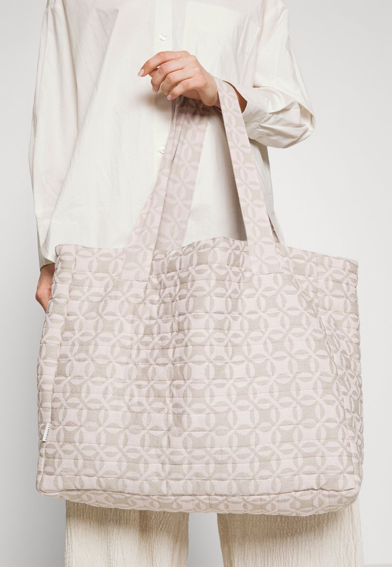 Personne portant une chemise blanche tient un grand sac cabas matelassé rose clair avec un motif géométrique en cercle et des poignées larges.