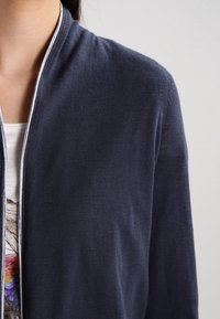 Tmavě modrý cardigan vyrobený z lehkého, strukturovaného materiálu. Disponuje předním zipem, kontrastním bílým lemem a výstřihem ve tvaru lodičky, vrstveným přes potiskovou blůzu.