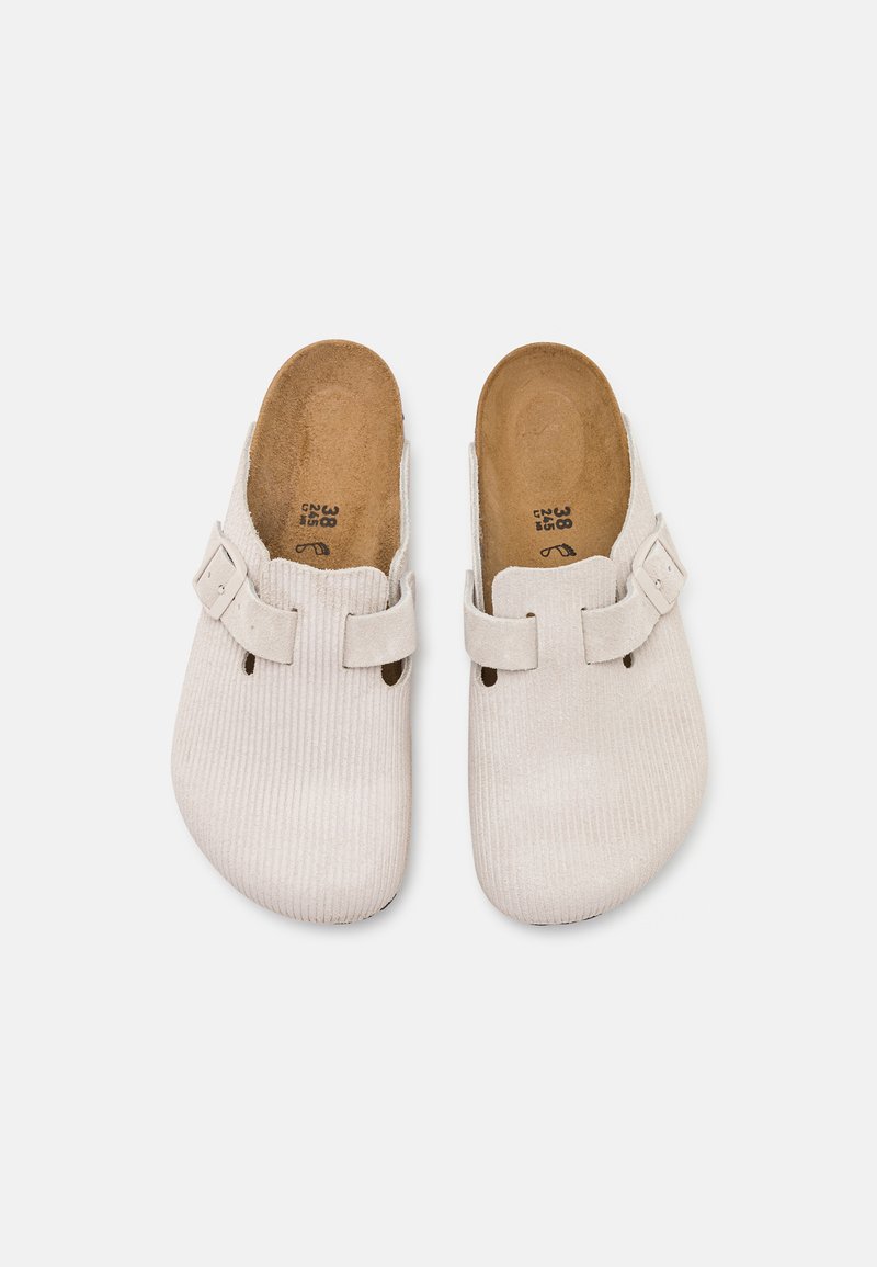 Zapatos de tela de pana blanca tipo slip-on con una textura suave, que presentan dos detalles de tira y un diseño de parte trasera abierta. Estilo minimalista y forma ergonómica.
