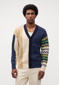 Multifarvet cardigan med marineblå, beige og creme sektioner, med teksturerede mønstre og V-hals. Inkluderer sorte knapper og levende accenter.