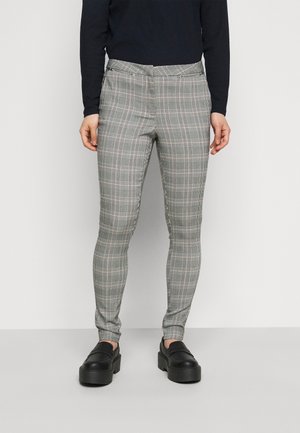Vrouw draagt grijze slim-fit geruite broek, zwarte top met lange mouwen en zwarte plateau loafers, staand voor een effen lichte achtergrond.