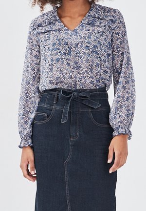 Femme portant un chemisier bleu à fleurs à manches longues, rentré dans une jupe en denim foncé taille haute avec une ceinture nouée à la taille.
