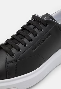 Fekete szintetikus bőr sportcipő fekete fűzőkkel, fehér talppal, dombornyomott "Armani Exchange" logóval és kontrasztos varrási részletekkel.