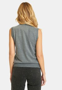 Suéter gris sin mangas con un patrón texturizado de pequeños puntos, cuello y dobladillo acanalados. Combinado con jeans oscuros que presentan acentos similares.
