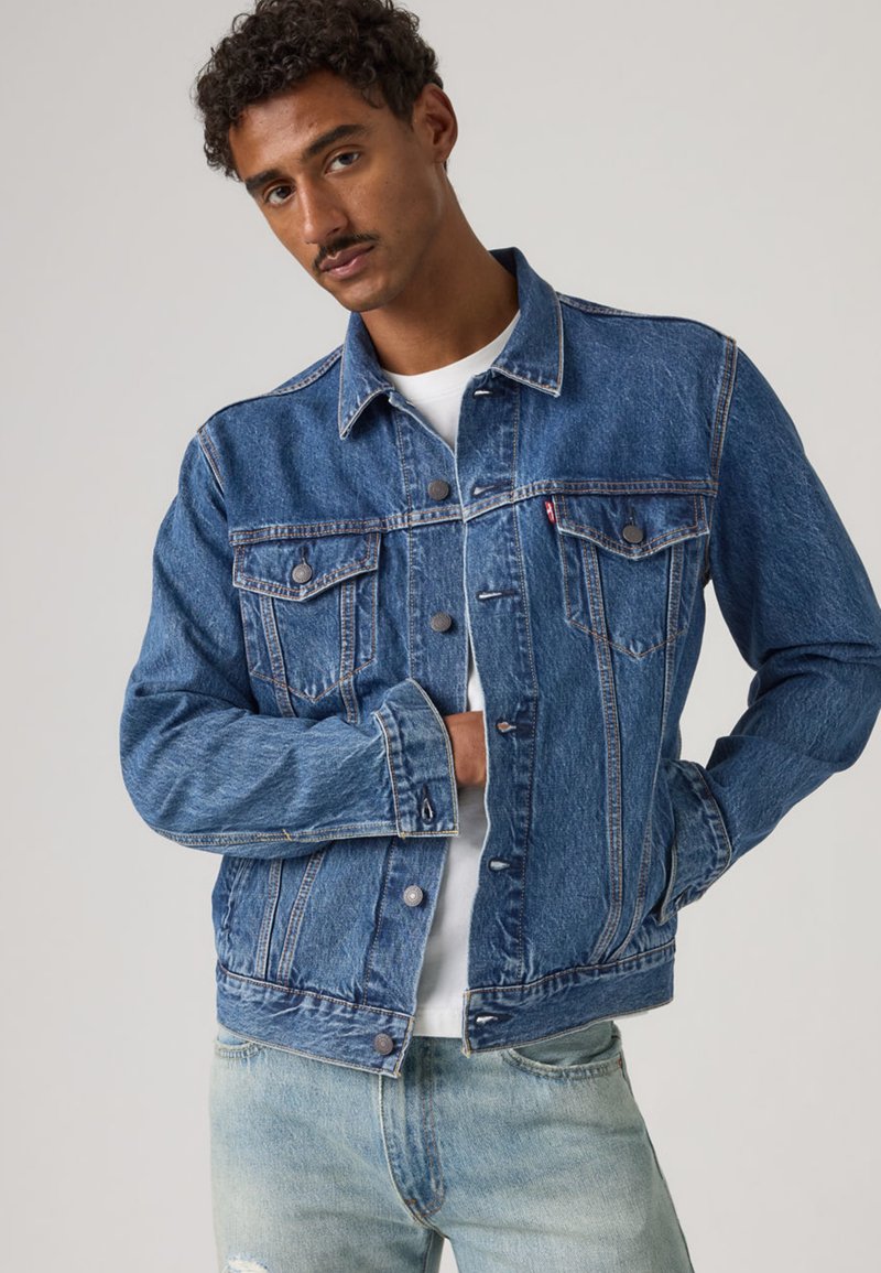 Blaue Jeansjacke mit klassischem Design, durchgeknöpft, zwei Brusttaschen und lockerer Passform, kombiniert mit hellen Jeans und einem weißen Hemd.