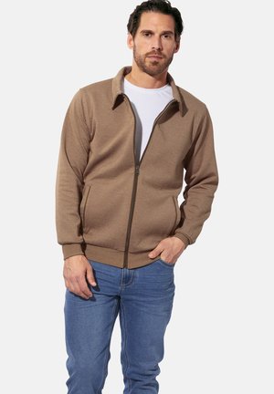 Man wearing a brown zip-up jacket over a white t-shirt and blue jeans, standing with one hand in pocket against a plain background.