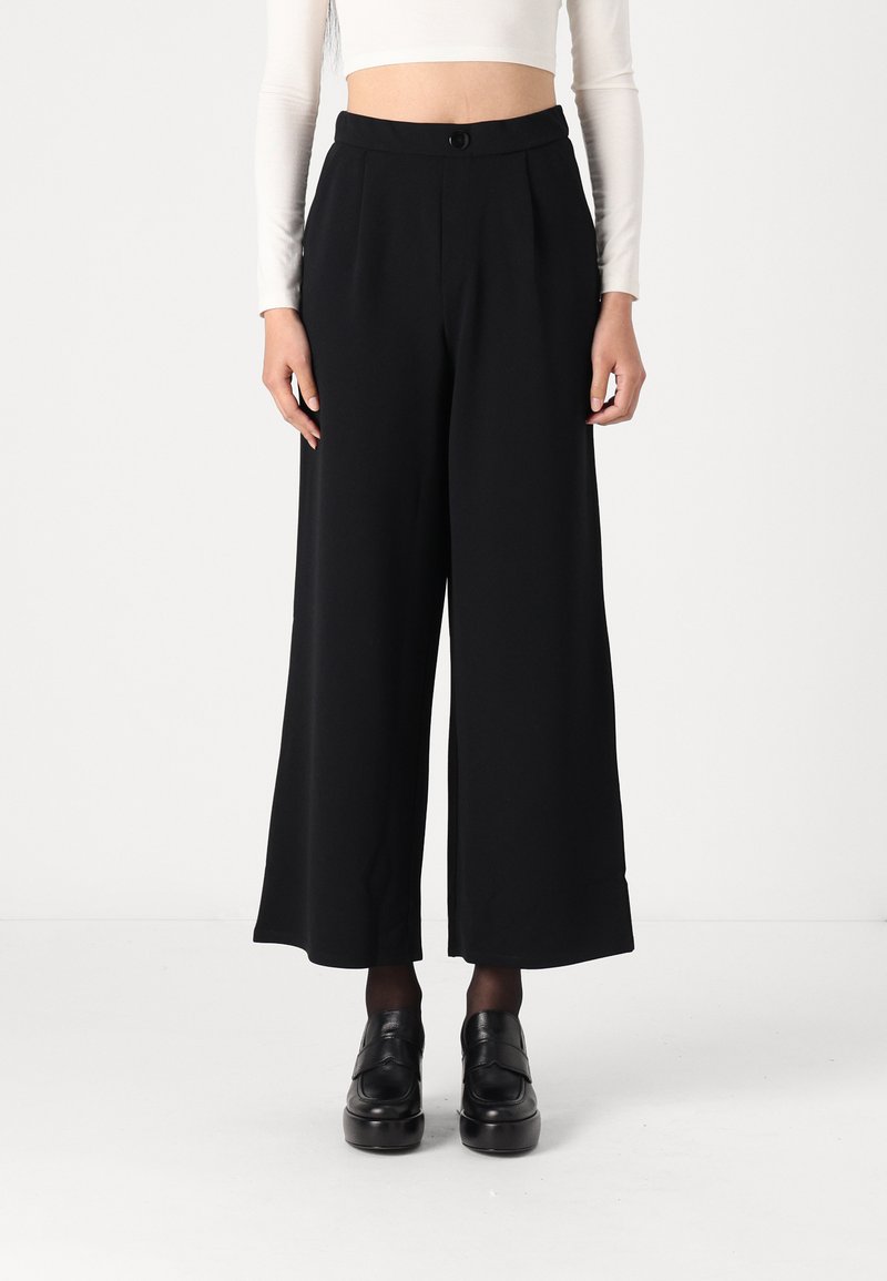 Black wide-leg trousers made from smooth fabric, featuring a high waist, front pleats, and a side button closure. Paired with black platform shoes.