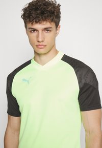 Camiseta atlética de manga corta en verde neón con acentos en negro y un patrón sutil en los hombros. Cuenta con un diseño de cuello tipo alto y un logo.