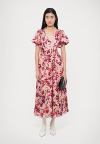 Vestito midi floreale rosa con maniche a sbuffo oversize, dotato di una cintura in vita e una gonna fluente. Indossato con scarpe bianche e una pochette nera.
