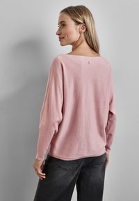 Pull rose clair, ample, avec un col large, des manches longues et une finition légèrement texturée. Il présente une petite étiquette à l'arrière.