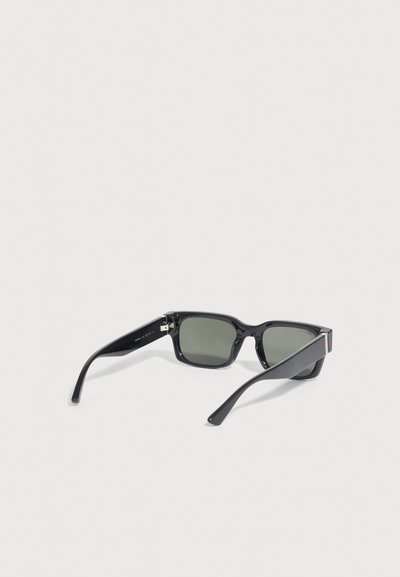 Μαύρα ορθογώνια γυαλιά ηλίου με σκοτεινούς φακούς και ελαφρώς κυρτά πλαϊνά, ανοιχτά τοποθετημένα σε ανοιχτό γκρι επιφάνεια.