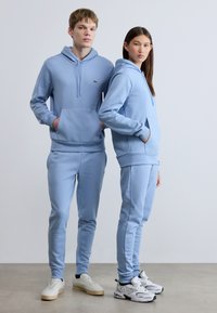 Σετ ανοιχτό μπλε φούτερ με κουκούλα και φόρμα, κατασκευασμένο από μαλακό ύφασμα. Διαθέτει τσέπη καγκουρό, ελαστικές μανσέτες και λεπτομέρεια λογότυπου στο στήθος και το μηρό.