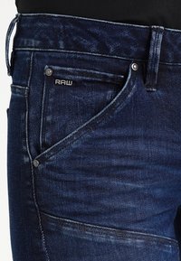 Mörkblå jeans i denim med en något urtvättning. Har metallbeslag och en loggetikett med texten "RAW" på framlommen.