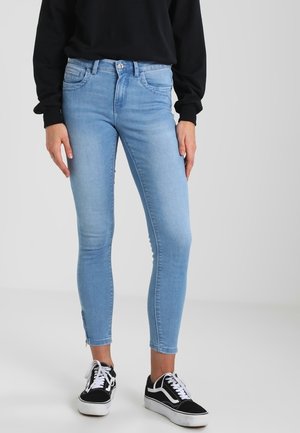 Person wearing light blue skinny jeans, black long-sleeve top, and black-and-white sneakers, standing against a plain background.