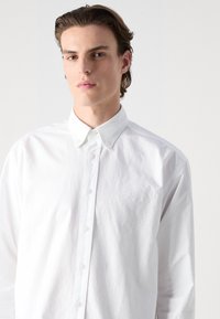 Biela košeľa s button-up zapínaním vyrobená z bavlny. Má klasický golier, dlhé rukávy a jemne vyšívané logo na hrudi. Hladká textúra.