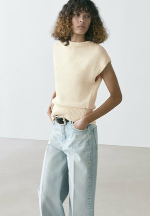 Frau mit lockigem Haar, die ein cremefarbenes, ärmelloses Strickoberteil und hellblaue Jeans mit weitem Bein trägt, mit Händen in den Taschen vor einem einfarbigen Hintergrund.