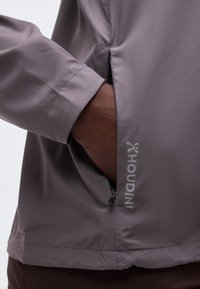 Χέρι που έχει τοποθετηθεί στην πλαϊνή τσέπη με φερμουάρ γκρι μπουφάν Houdini, δείχνοντας λεπτομέρεια του φερμουάρ και υφή υφάσματος.