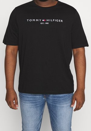 Hombre con camiseta negra de Tommy Hilfiger con logo y texto "EST. 1985", combinada con vaqueros azules desgastados, frente a un fondo liso.