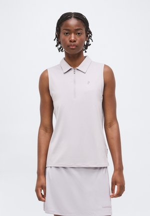 Žena nosi svetlosivé bezrukávové polo na zips a zhodnú sukňu, stojaca na jednoliatom bielom pozadí.