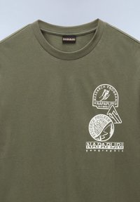 Camiseta de algodón verde oliva con cuello redondo, que presenta logotipos y gráficos impresos en blanco en la parte delantera. Textura suave, diseño casual.