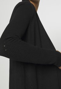 Cárdigan negro de punto con mangas largas de textura acanalada y un ajuste holgado, que presenta un sutil detalle en los extremos de las mangas.