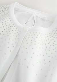 Valkoinen neuletakki hopeanvärisillä helmikoristeilla, nappisuljenta ja pehmeä tekstuuri. Kauluksessa pieni rusetti, merkissä näkyy merkki ja alkuperä.