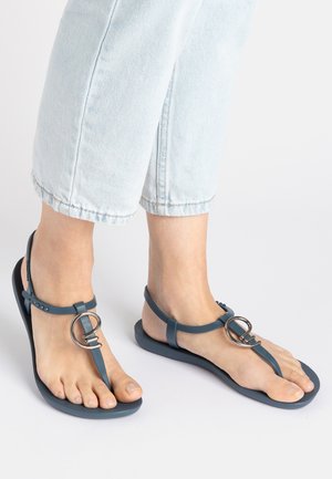 Blue sandal with a T-strap design, featuring a metal circular accent and textured sole. Made of flexible material; flat profile.