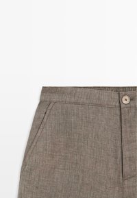 Close-up van bruine, gestructureerde stoffen broek met zijzak, knoopsluiting en gedeeltelijk elastische tailleband op witte achtergrond.