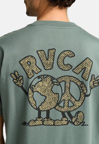 T-shirt in cotone verde con una grafica nera e dorata della Terra con segno di pace. Il design include caratteri giocosi e lettering audace.