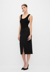 Bodysuit noir côtelé avec larges bretelles, associé à une jupe midi noire avec une fente à l'avant. Modèle portant des escarpins noirs à bout pointu.