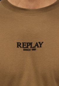 Tissu marron avec texte noir "REPLAY SINCE 1981" imprimé sur la zone de la poitrine d'un vêtement à col rond.