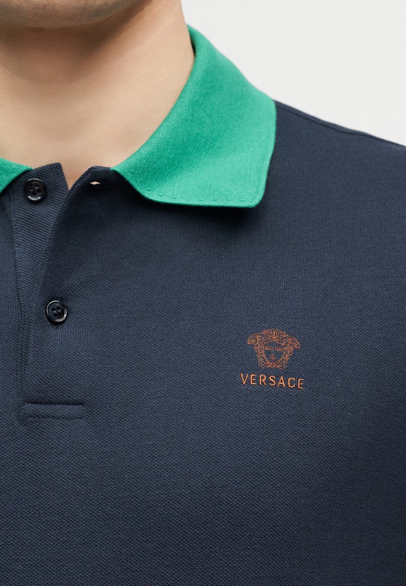 Man draagt een marineblauw poloshirt met een groene kraag, met een oranje Versace-logo op de linkerborst.