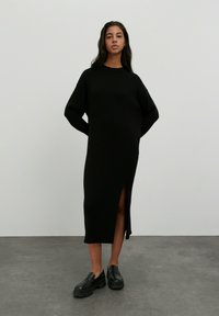 Schwarzes geripptes Strickkleid mit langer Silhouette und seitlichem Schlitz, das eine lockere Passform und Überschulter-Ärmel aufweist. Getragen mit schwarzen Plateau-Schuhen.