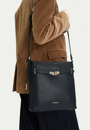 Persona che indossa una giacca di velluto a coste marrone con una borsa a tracolla in pelle nera con chiusura dorata e logo LASOCKI.