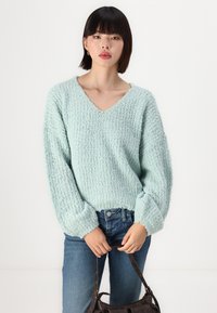 Pull vert menthe clair en col en V avec un tissu texturé, des manches volumineuses et des poignets côtelés, associé à un jean bleu foncé et un sac à main marron.