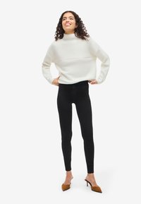 Bela rebrasta pletena jopica z visokim ovratnikom, v kombinaciji s črnimi ozkimi kavbojkami in rjavimi čevlji s koničastimi prsti. Model stoji samozavestno.