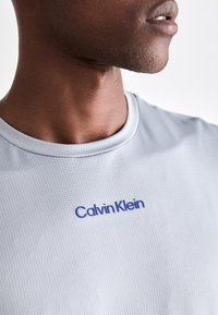 Cămășuță sport cu mânecă scurtă de culoare gri deschis, realizată dintr-un material texturat. Prezintă un logo albastru „Calvin Klein” pe piept. Guler rotund.