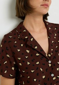 Chemise marron à manches courtes avec un imprimé léopard noir et beige, avec un col à revers et des détails de boutons. Texture lisse.