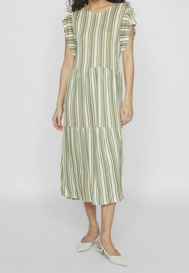 Femme portant une robe midi à rayures verticales vertes et blanches avec de courtes manches volantées, associée à des escarpins blancs à bout pointu et bride arrière.