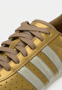 Zlatá kožená teniska s béžovými šnúrkami, striebornými pruhmi, perforovanými detailmi a textúrovaným povrchom. Zlaté akcenty na očkách šnurovania.