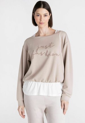Jonge vrouw met stijl donker haar draagt een beige sweatshirt met "just fashion" parelletters, een witte gelaagde zoom en lichte geribbelde broek.