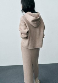 Beige pletená mikina s kapucí a stahovací šňůrkou, volnější střih, s žebrovaným spodním lemem, v kombinaci s odpovídající sukýrkou po kolena s rozparkem na boku.