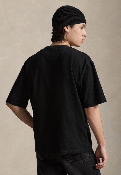 Νεαρός άνδρας με μαύρο σκούφο και φαρδιά μαύρη μπλούζα, φωτογραφημένος από πίσω σε ένα απλό μπεζ φόντο.