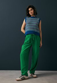 Gebreide blauwe mouwloze top met multi-kleurige strepen, gecombineerd met losse groene broek en beige sneakers met zwarte strepen.