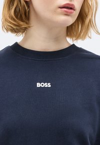 Νέος με κοντά ξανθά μαλλιά, φορεί μπλε κατάδυμα με τη λέξη "BOSS" σε λευκό στο στήθος.