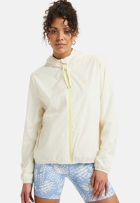 Lightweight, cream-colored zip-up jacket with a hood and elastic cuffs, featuring a subtle logo and yellow zipper. Paired with blue patterned shorts.