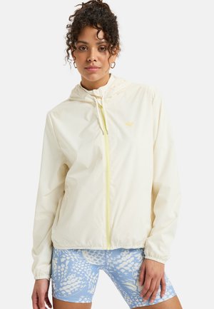 Lightweight, cream-colored zip-up jacket with a hood and elastic cuffs, featuring a subtle logo and yellow zipper. Paired with blue patterned shorts.