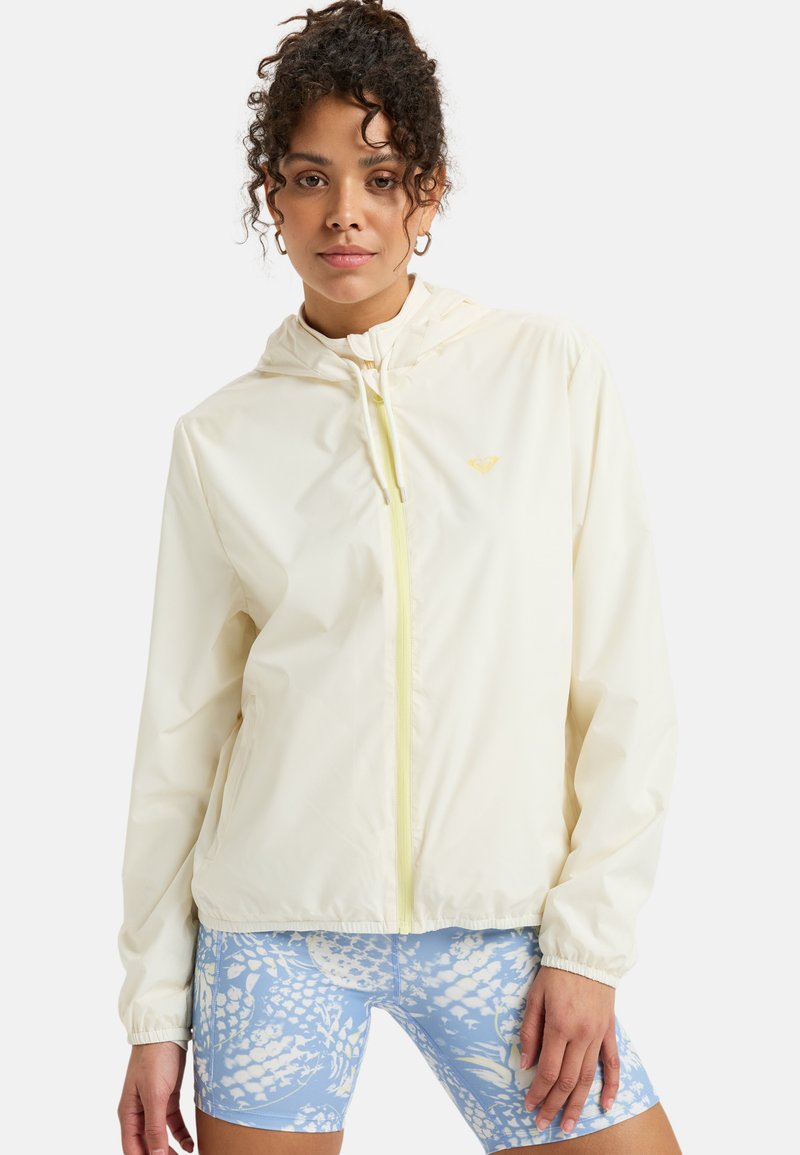 Lightweight, cream-colored zip-up jacket with a hood and elastic cuffs, featuring a subtle logo and yellow zipper. Paired with blue patterned shorts.