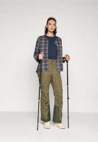Geruite shirt in groen, blauw en bordeauxrood patroon over een marineblauw t-shirt; olijfkleurige cargo-broek met groene panelen; houdt trekkingstokken vast.